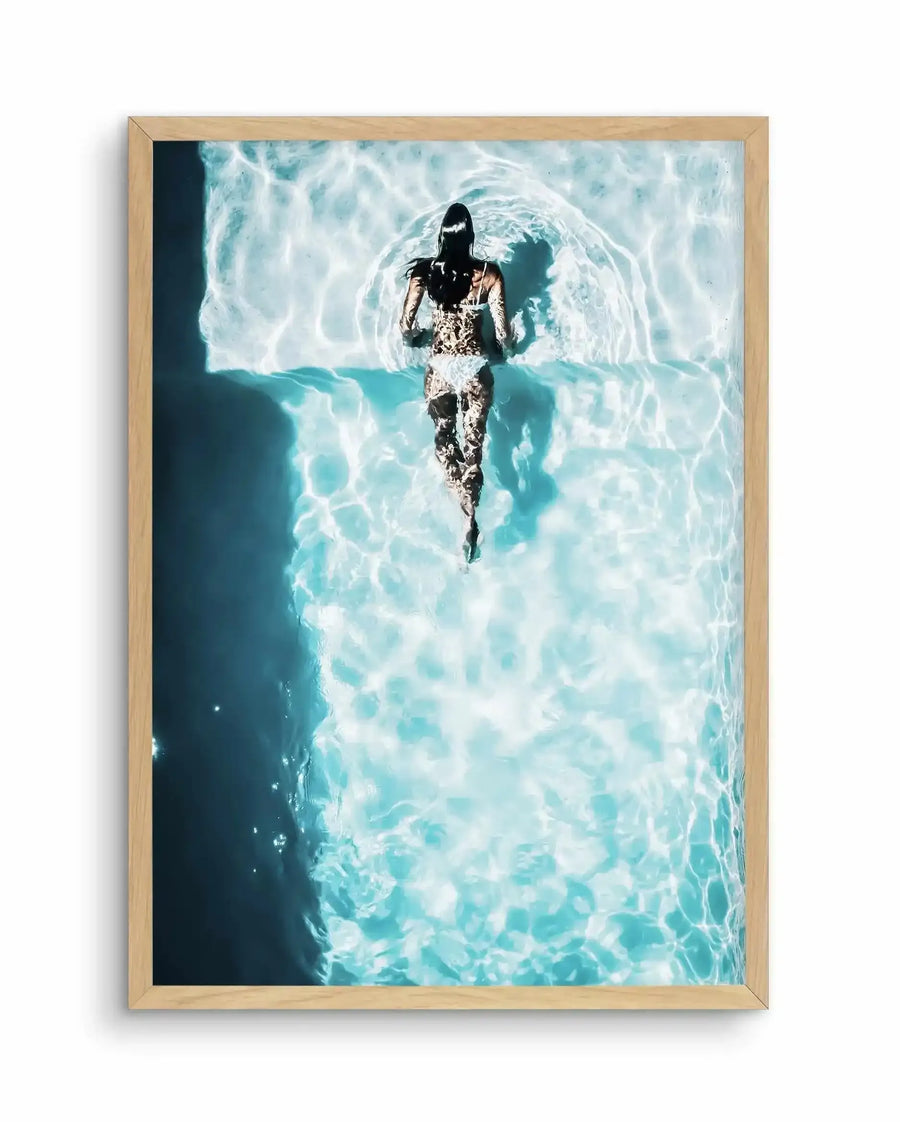 Aerial photograph of a woman in a white lace swimsuit floating in crystal-clear turquoise water, viewed from directly above. The water's surface shows rippling patterns and light refraction in shades of aquamarine, white, and pale blue. The woman's dark hair contrasts against the bright water. The photograph is displayed in a natural wood frame with a vertical portrait orientation, creating a serene, summery composition.