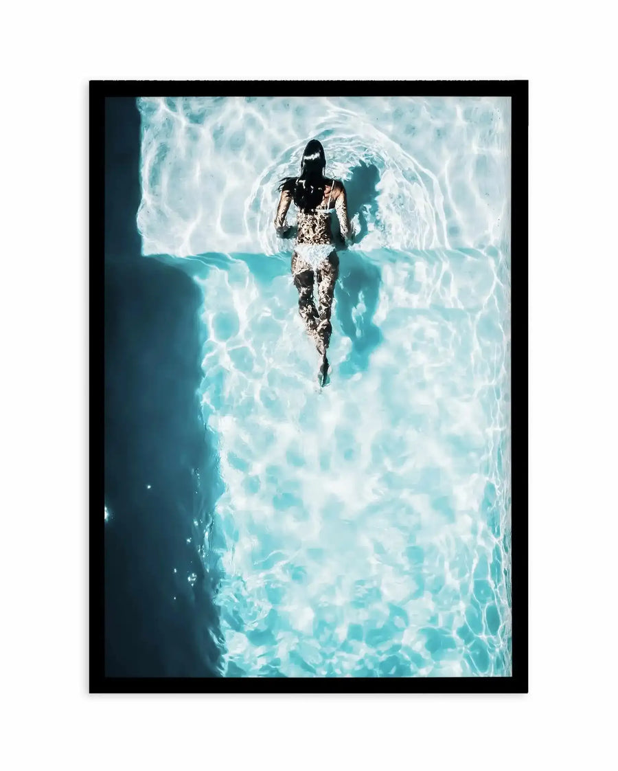 Aerial photograph art print in black frame showing a woman viewed from directly above, standing in shallow turquoise water. The woman wears a white lace swimsuit and has long dark hair. The water below creates rippling patterns in shades of turquoise, white, and pale blue, with sunlight refracting through creating luminous wave textures across the entire composition. The print has a serene, summery mood with high contrast between the figure and the reflective water surface.