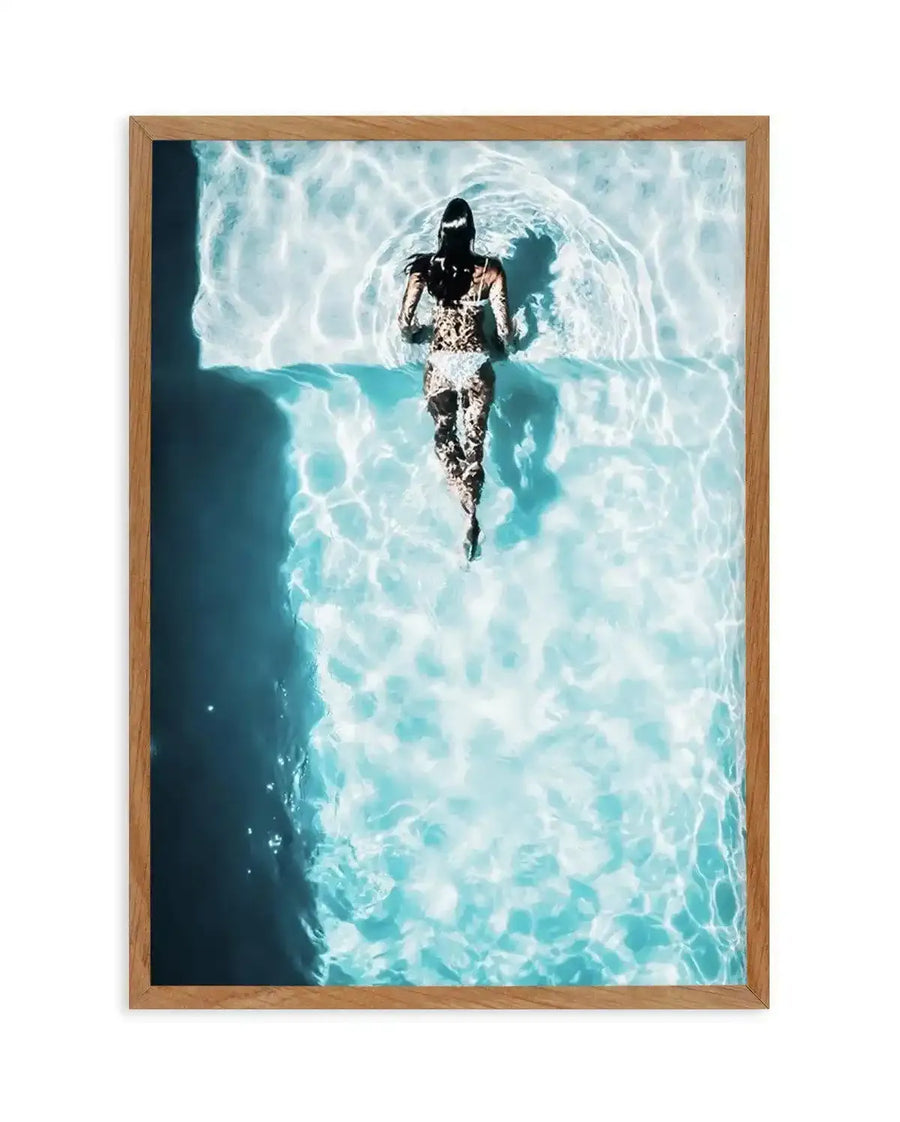 Aerial photograph in natural wood frame showing a person in a white lace dress standing in shallow turquoise ocean water, photographed from directly above. The water features bright white foam and rippling patterns in shades of aqua and teal. The person's silhouette appears centered against the luminous water backdrop, creating a striking contrast. The photograph has a serene, summery aesthetic with cool tones dominating the composition.