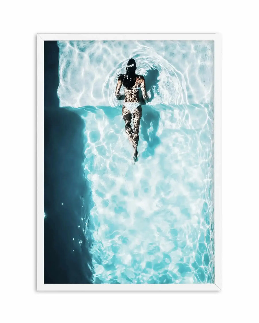 Framed aerial photograph of a woman swimming in a bright blue pool with a white border. The woman is seen from behind, wearing a light-colored, possibly leopard print, swimsuit, and her dark hair is wet and flowing. The water is a vibrant turquoise with strong sunlight creating shimmering, rippling patterns and reflections across the surface, and the pool wall on the left is a darker blue, indicating depth.