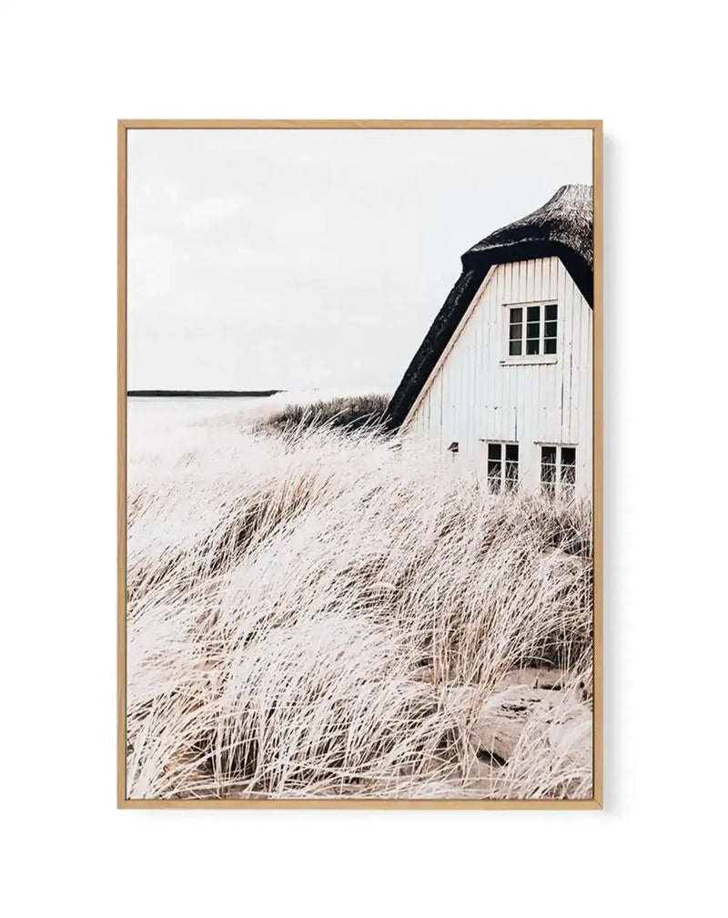 Framed canvas art print featuring a coastal landscape photograph. A white-painted wooden cottage with a dark thatched roof sits on a sand dune, with small rectangular windows visible on its front facade. Dried beach grasses in pale tan and cream tones cover the foreground and surrounding dunes. A calm body of water appears in the soft, overcast background. The photograph is mounted in a natural wood frame with a white border, creating a serene, minimalist coastal scene.