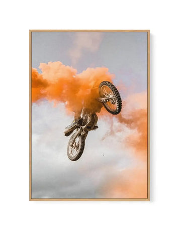 Framed canvas print of a motocross rider mid-air, surrounded by a large cloud of vibrant orange smoke against a pale grey and white sky. The rider is angled upwards with the front wheel of the dirt bike pointing towards the ground, capturing a dynamic moment of action. The print is displayed in a natural wood frame.