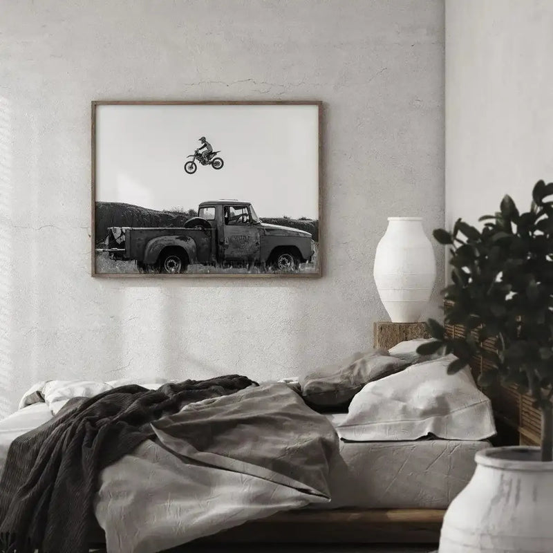 Large framed black and white art print displayed above a bed in a minimalist bedroom. The print features an old, rusty pickup truck parked in a field with a motocross rider mid-air on a dirt bike above it, set against a bright white sky. The print has a natural wood frame and white border. The bed is covered in neutral-toned linen bedding and a dark grey throw, with a white textured vase on a wooden block beside it and a potted plant in the foreground.