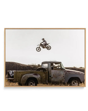 Framed canvas art print featuring a dynamic outdoor scene with a motocross rider mid-air above an old, rusty pickup truck. The rider, wearing a helmet and gear, is silhouetted against a bright, clear sky, with the dirt bike's wheels clearly visible. Below, the weathered, faded green and grey truck, with "HUNT" painted on its side, is parked in dry, grassy terrain, with a dark, rocky hill in the background. The print is displayed in a natural wood frame.
