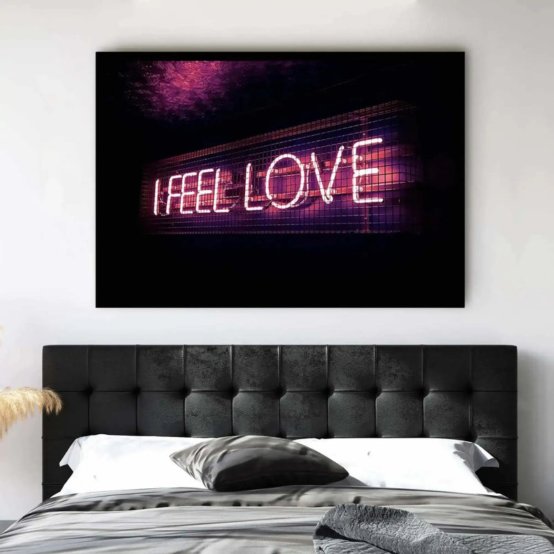 Lifestyle shot of a bedroom featuring a large art print with the words "I FEEL LOVE" in white neon tubing against a dark background with hints of purple and pink light. The print is displayed above a black upholstered headboard with square tufting, on a bed with white pillows, grey bedding, and a grey textured throw. Dried pampas grass is visible on the left side of the headboard.