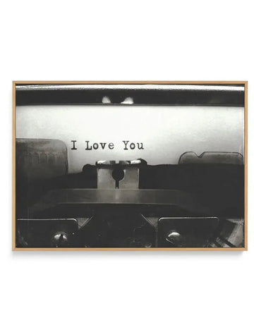 Black and white photograph of a typewriter shot from above, showing the paper roller and keys in sharp detail. The typed text "I Love You" appears on white paper in the center. The image has high contrast with deep blacks and bright whites, creating a vintage aesthetic. The photograph is mounted on a canvas print and housed in a natural wood frame with a white border.