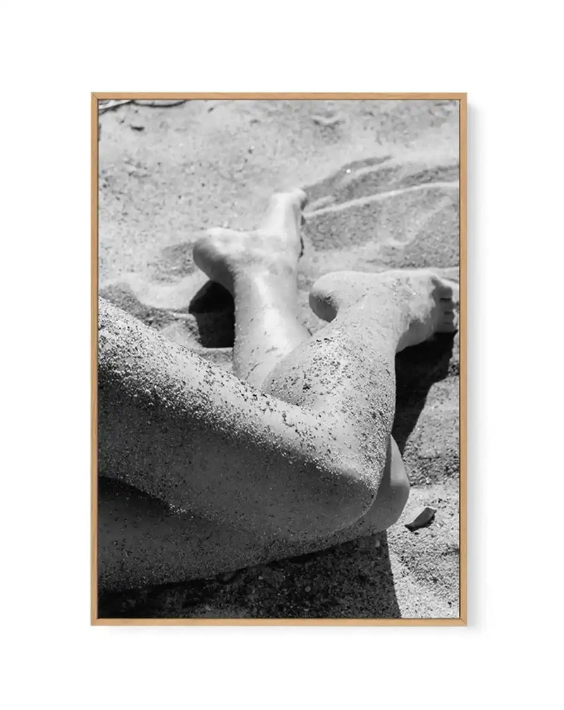 Black and white photograph of weathered stone sculptures or architectural fragments partially buried in sand, displayed in a natural wood frame. The image shows carved stone forms with curved surfaces and hollow openings, creating strong shadows and textural contrast. The grainy quality and monochromatic palette emphasize the tactile, eroded surface details of the ancient or aged stone pieces. White border frames the photograph within the wooden frame.
