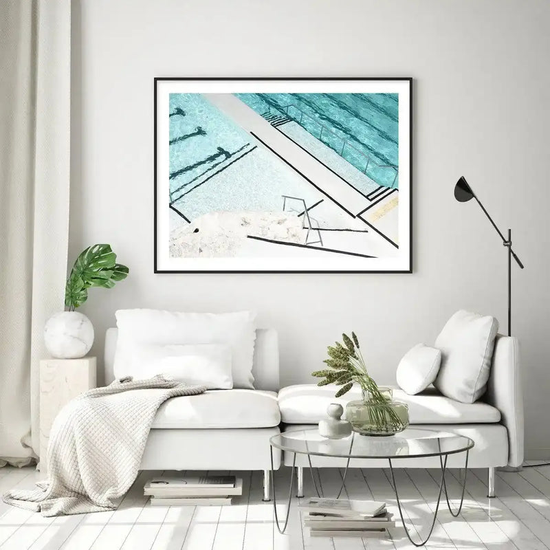 Large framed art print displayed above a white sofa in a minimalist living room. The print features an aerial photograph of the iconic Bondi Icebergs pool, with distinct lane lines and clear turquoise water, captured in a modern, graphic style with a black frame and white border. The room includes a textured cream throw, a monstera plant in a marble vase, and a glass coffee table with dried grasses.