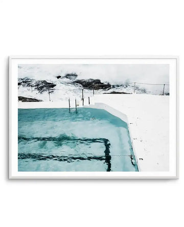 Framed art print featuring an aerial photograph of an icy geothermal pool with turquoise water surrounded by white snow and black volcanic rock. A wooden ladder extends into the clear water, with distant snowy mountains and suspension bridge visible in the background. The image has a cool, minimalist aesthetic with soft whites, teals, and charcoal tones. Presented in a slim silver frame with white border.