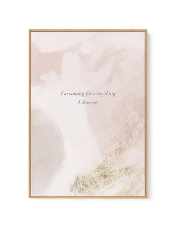 Framed canvas art print with a natural wood frame featuring an abstract landscape photograph in muted beige and cream tones. Snow-covered mountainside or terrain visible in the lower right corner with soft, blurred peaks in the background. Centered in italicized serif font is the affirmation text "I'm coming for everything I deserve" in dark gray. The overall composition has a serene, minimalist aesthetic with a soft, dreamy quality enhanced by the neutral color palette and photographic texture.
