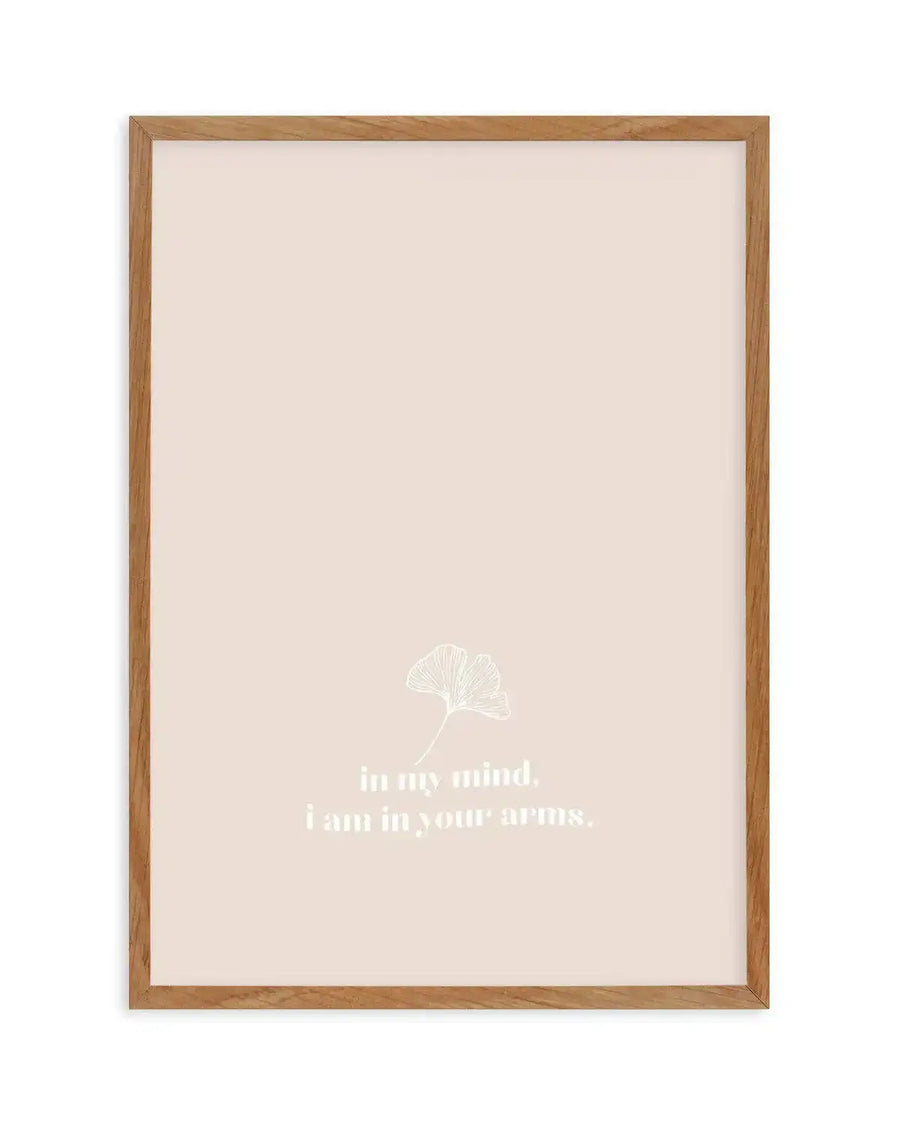 Framed art print with natural wood frame displaying a minimalist design on a soft beige background. A delicate white line drawing of a ginkgo leaf appears in the center-upper portion. Below it, white serif text reads "in my mind. i am in your arms." in two lines. The print has a white border and is mounted in a light oak-toned wooden frame.