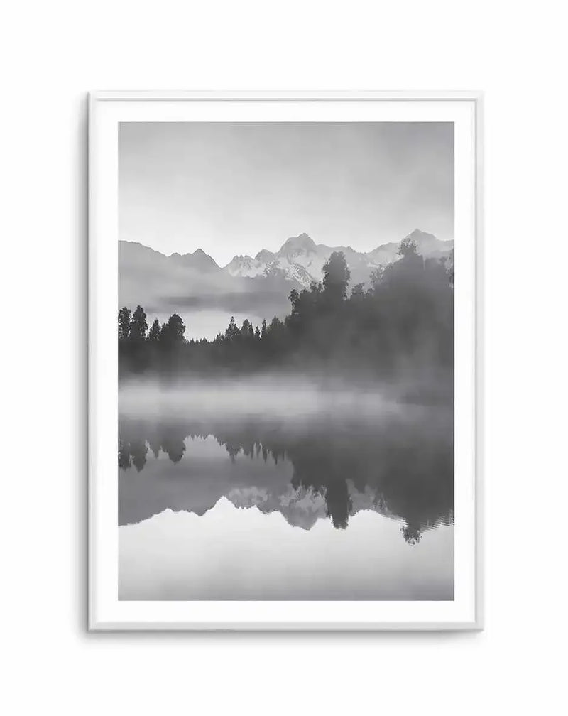 Framed black and white photograph of a serene mountain and lake landscape with a white frame and white border. The composition features snow-capped mountains in the distance, a dense forest along the middle ground, and a misty lake in the foreground with a clear reflection of the mountains and trees.