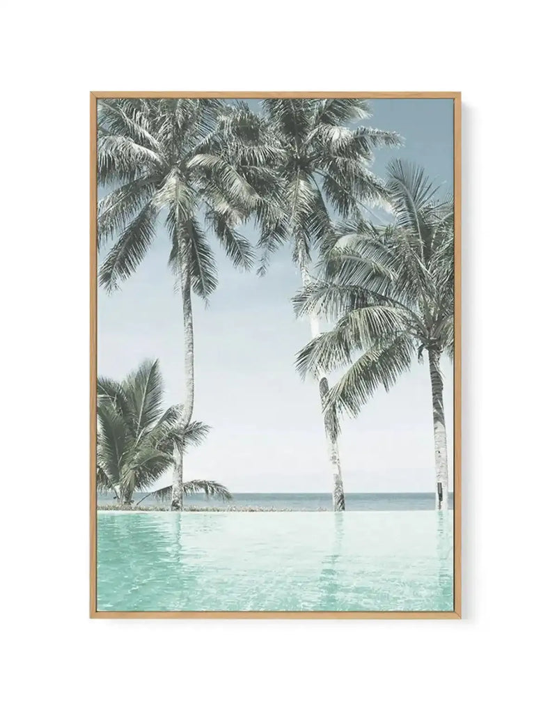Framed canvas art print featuring a serene poolside scene with tall coconut palm trees standing in shallow turquoise water. Clear blue sky visible between the fronds. Ocean and distant shoreline visible in the background. Natural wood frame with white border. Photograph-style image with soft, calm color palette of aqua, seafoam green, pale blue, and cream tones.