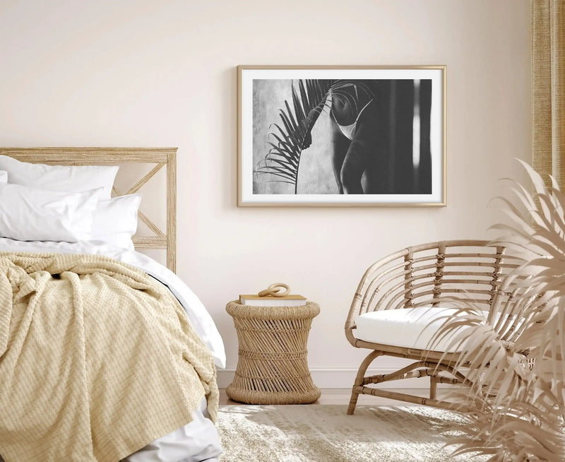 Framed black and white photography art print displayed above a rattan side table in a minimalist bedroom. The print features a person in a white bikini top holding a large palm frond, with soft light and shadow creating a serene mood. The room includes a wooden bed frame with white bedding and a cream textured throw, a rattan armchair, and dried palm fronds in the foreground.