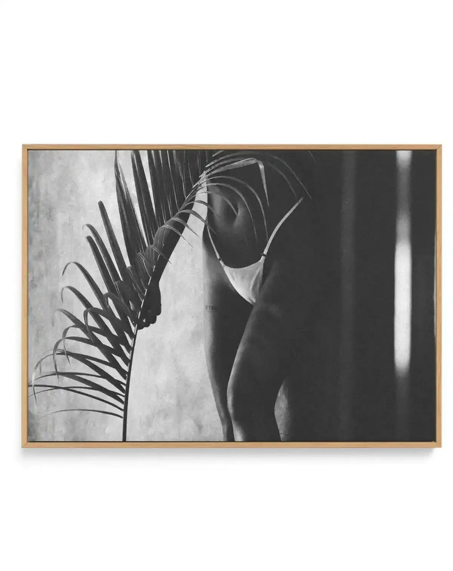 Framed black and white photograph of a person in a bikini holding a large palm frond, printed on canvas with a natural wood frame. The composition shows the person's torso and legs, partially obscured by the dark frond, against a textured light background on the left and a dark, shadowed background on the right with a vertical stripe of light.
