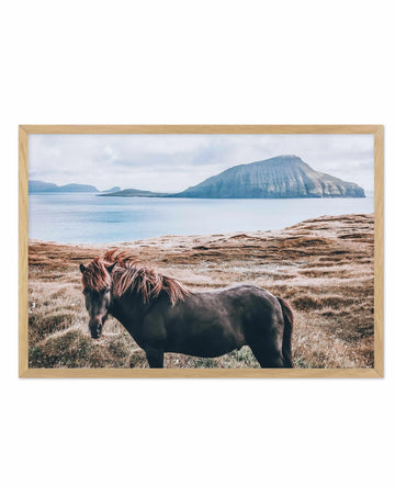 Framed art print featuring a dark brown Icelandic horse with long reddish-brown mane standing in sparse, tan grassland facing left. Behind the horse stretches a calm turquoise sea with a distinctive cone-shaped volcanic island with vertical columnar formations visible across the water. Smaller islands and mountains appear in the misty background under an overcast sky. The photograph is mounted in a natural wood frame with white border.