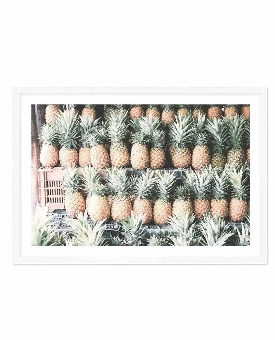 Framed photography art print of a market stall filled with rows of pineapples, displayed in a white frame with white border. The composition features three rows of ripe pineapples with green leafy tops, creating a textured pattern of golden-brown and green.