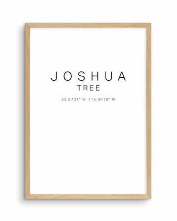 Joshua Tree Coordinates Art Print in natural wood frame. The print features minimalist black text on a white background reading "JOSHUA TREE" with coordinates "33.8734° N, 115.9010° W" displayed below in a smaller sans-serif font. The artwork is centered on the page with ample white space. The frame is light natural wood with a simple rectangular border, displayed against a white background.