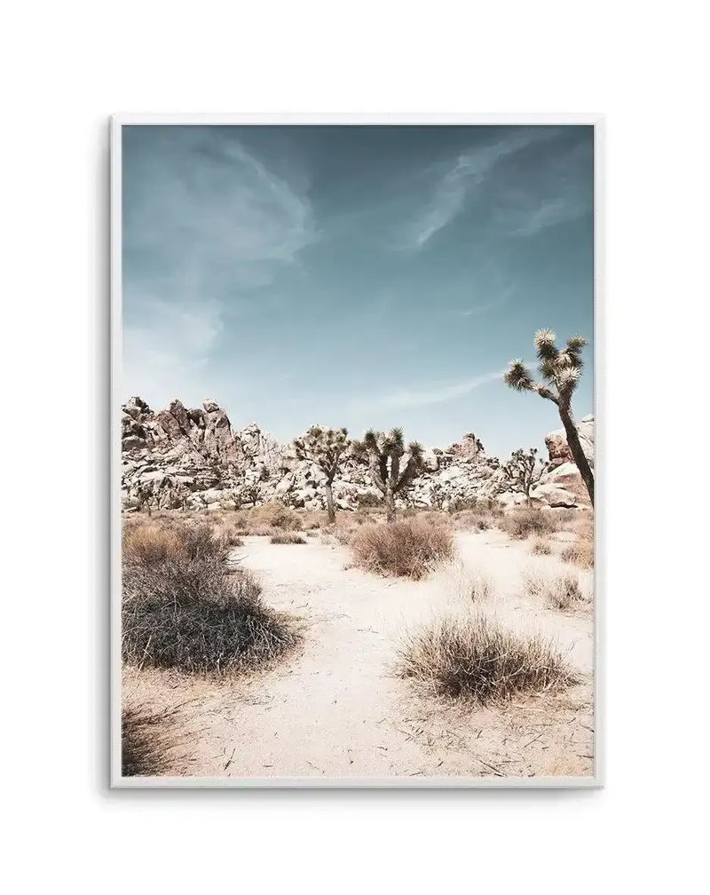 Framed photography art print of a desert landscape featuring Joshua trees and rock formations under a pale blue sky with wispy clouds, displayed in a white frame with a white border. The composition captures the sandy ground with sparse dry bushes in the foreground, leading to a cluster of Joshua trees and large, light-colored boulders in the midground, with a single Joshua tree on the right.