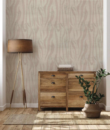 Jungle Stripes Wallpaper featuring a repeating vertical striped pattern in soft taupe, blush pink, and cream tones with organic, wavy line details creating a textured, natural appearance. The wallpaper is displayed on a bedroom wall behind a wooden dresser with metal cup handles, a tripod floor lamp with brown linen shade, a potted green plant in a woven basket, and a natural fiber area rug, demonstrating the wallpaper's sophisticated, neutral aesthetic in an interior setting.