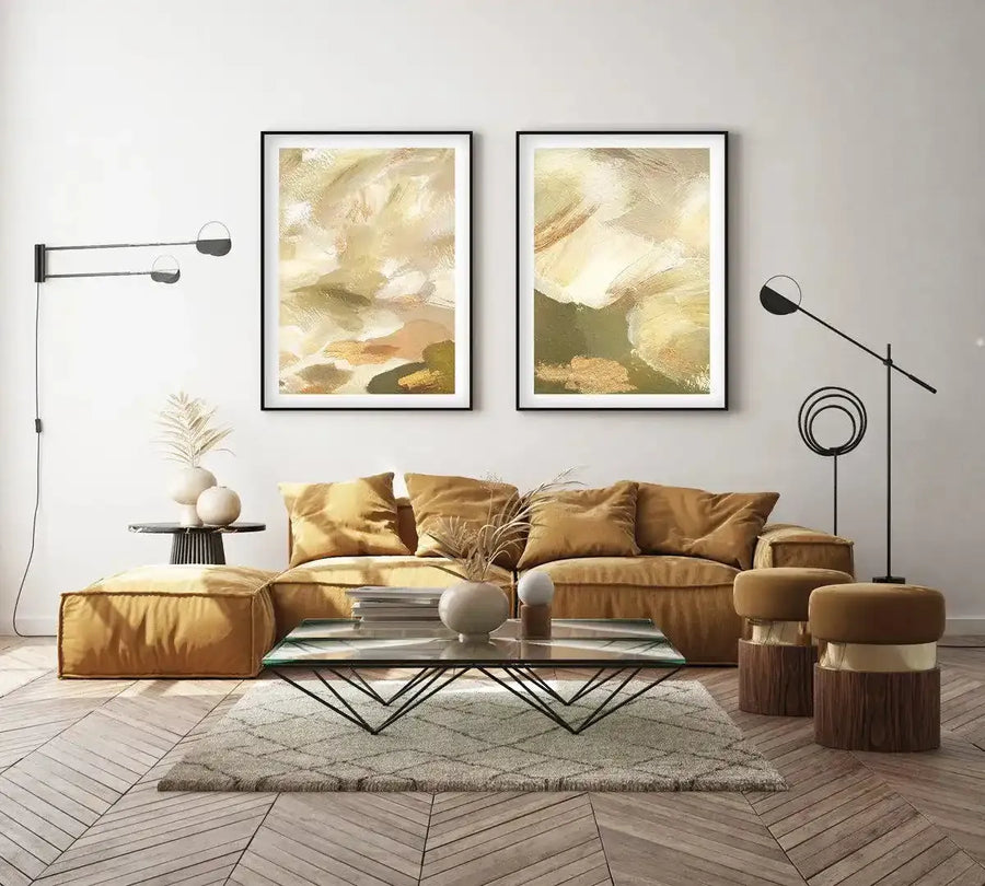 Khaki Luxe I Art Print displayed in a black frame with white border, hanging on a light gray wall above a gold sectional sofa. The artwork features an abstract composition of soft, layered brushstrokes in cream, sage green, warm gold, and earthy tan tones, creating an organic, painterly effect reminiscent of natural landscapes or geological formations. The print is shown in a styled living room setting with modern furnishings, complementing the room's neutral and warm color palette.