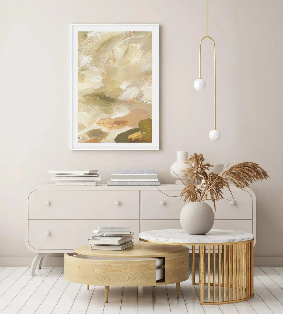 Abstract art print featuring broad, textured brushstrokes in a palette of warm khaki, soft cream, muted sage green, and hints of shimmering gold, creating a layered, organic composition suggestive of a natural landscape. The print is displayed in a simple white frame with a white border, hanging above a light beige dresser in a minimalist living space.