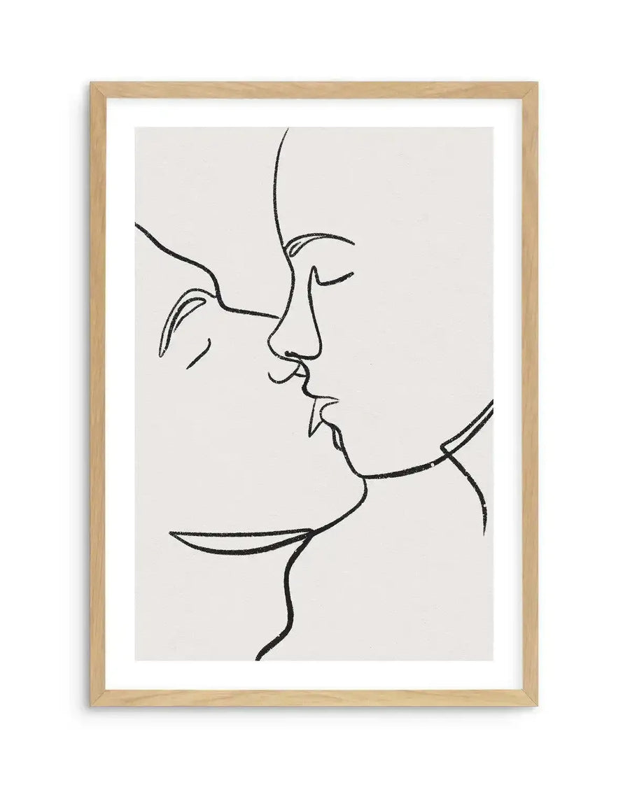 Line art print featuring two faces in profile, almost touching in a kiss, rendered in a single continuous black line on a cream background, printed on fine art paper and displayed in a natural oak frame with white border.