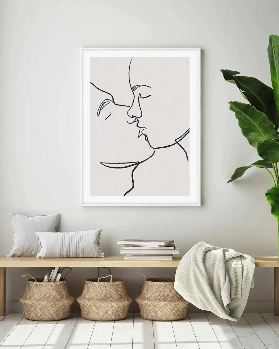 Large framed art print displayed above a wooden bench in a minimalist living room. The print features a continuous line art illustration of two faces kissing, rendered in black on a light grey background, with a white frame and white border. The room includes two white cushions, a stack of books, a cream throw blanket, and three woven baskets on a white wooden floor.