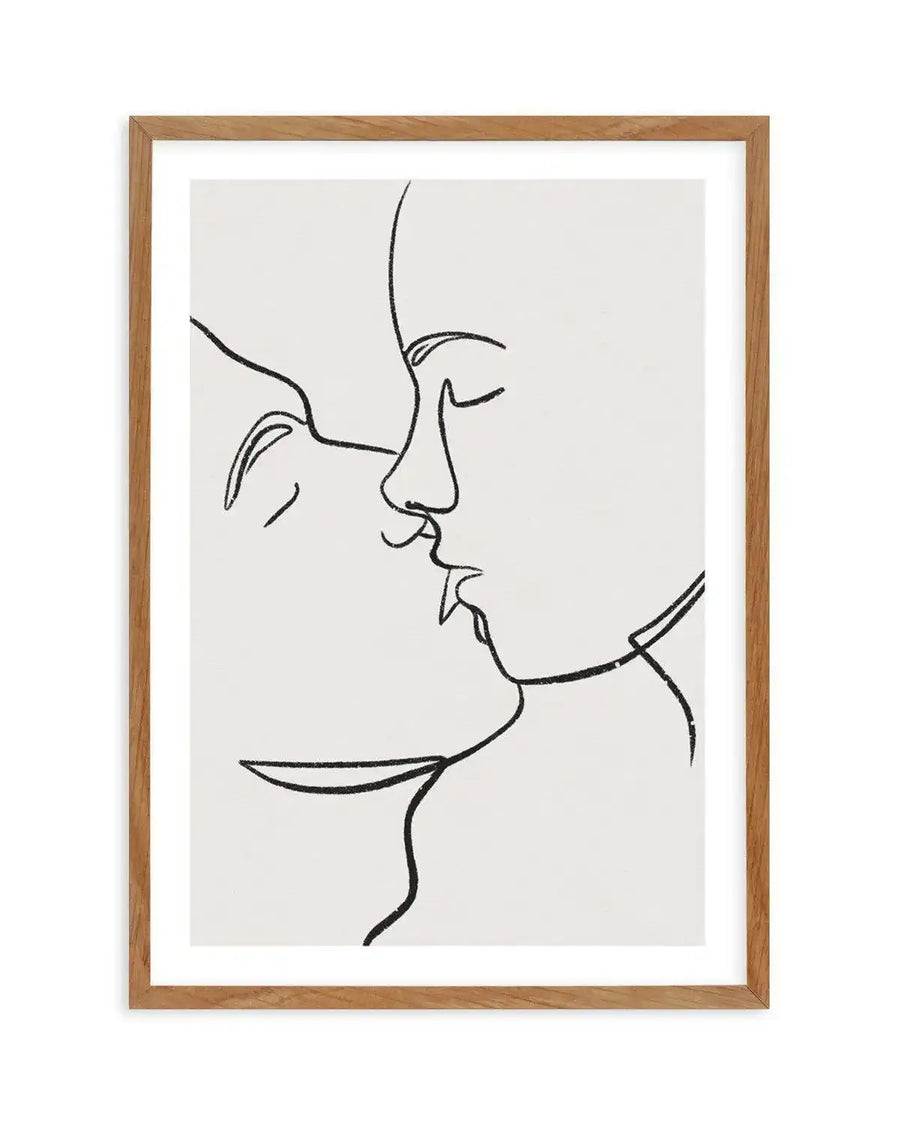 Line art print featuring two faces in profile, leaning in for a kiss, drawn with a single continuous black line on a white background, displayed in a natural oak frame with white border. The faces are minimalist, with closed eyes, and the lines have a slightly textured, charcoal-like quality.