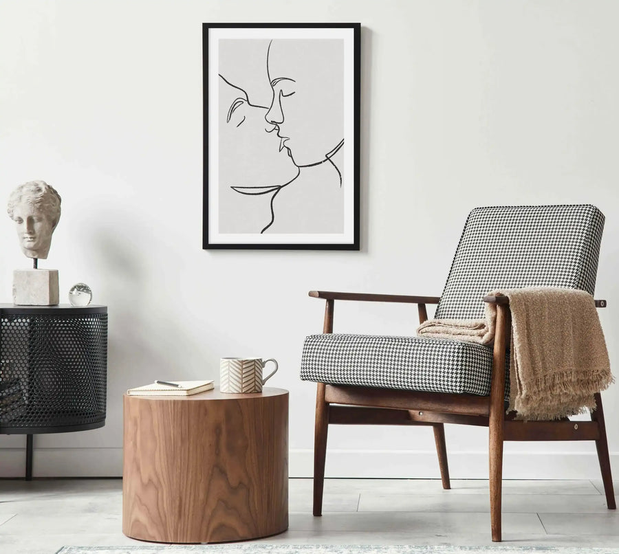 Large framed art print displayed above a round wooden side table in a minimalist living room. The print features a black and white line art illustration of two faces in profile, kissing, with a black frame and white border. The room includes a houndstooth patterned armchair with a beige throw, a black perforated metal cabinet with a classical bust and glass paperweight, and a patterned rug on a light grey floor.