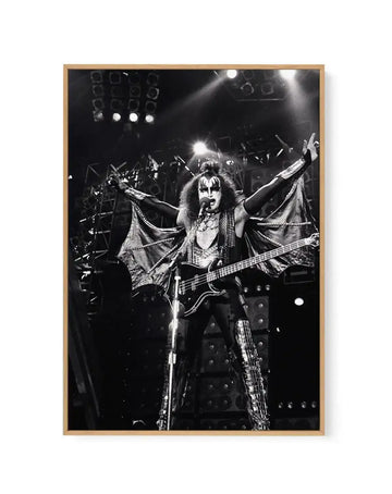 Black and white framed canvas art print featuring a dramatic live performance photograph of a rock musician with face paint and long hair, wearing an ornate studded costume with wing-like fabric elements, playing bass guitar on stage with arms raised. Stage lighting and amplifiers visible in the background. Image mounted in natural wood frame with white border.