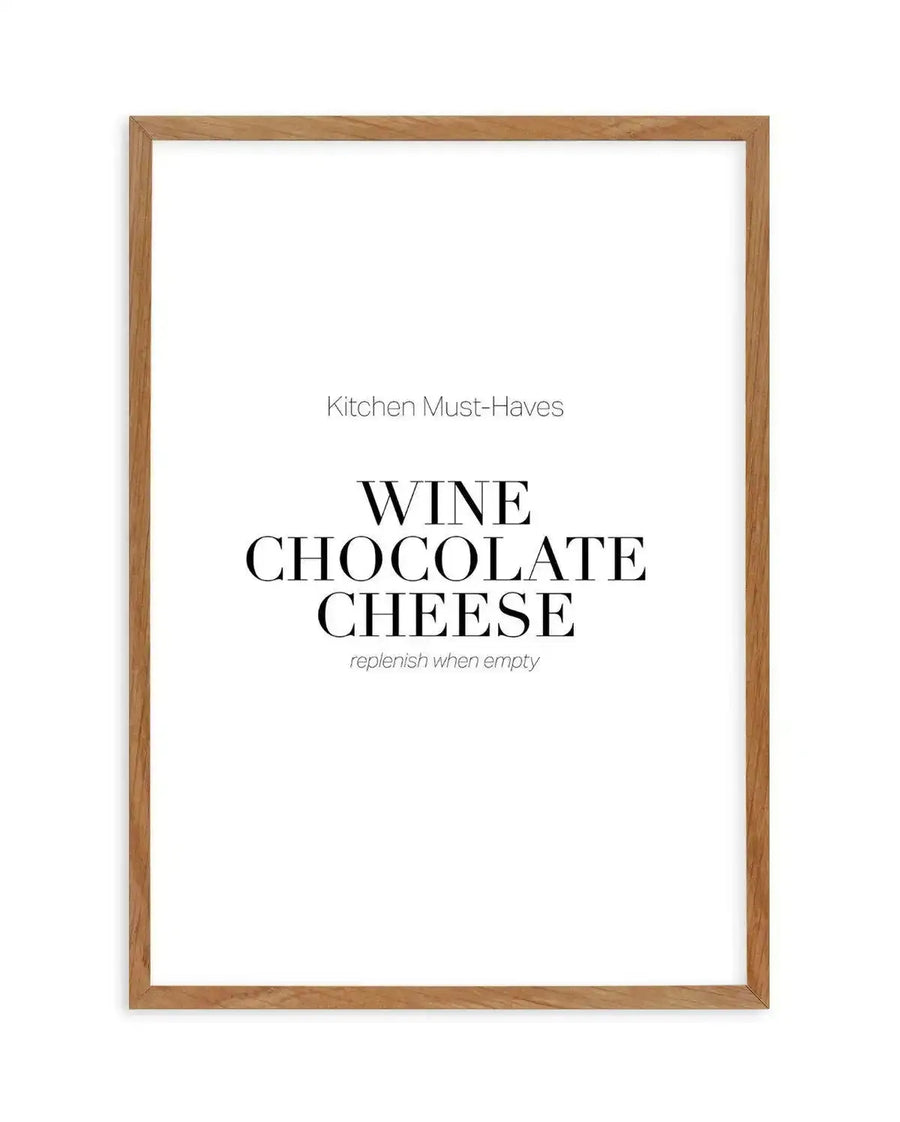 Framed art print with natural wood frame displaying text-based design. The print reads "Kitchen Must-Haves" in small gray sans-serif at top, followed by "WINE CHOCOLATE CHEESE" in large bold black serif letters in the center, with "replenish when empty" in small gray italic text below. White background with clean, minimalist composition. Natural wood frame with visible grain texture.