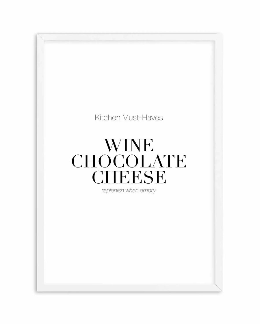 White framed art print featuring centered typography on a white background. The text reads "Kitchen Must-Haves" in thin, light gray sans-serif font at the top, followed by "WINE CHOCOLATE CHEESE" in large black serif capitals, and "replenish when empty" in small italic gray text below. The print is mounted in a white wooden frame with a simple, clean border.