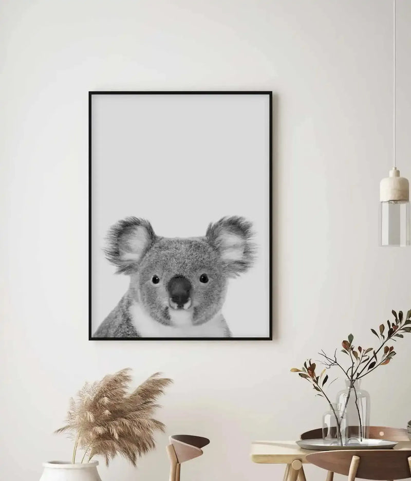 Black-framed art print of a koala's face in black and white photography, displayed on a white wall above a wooden table styled with dried pampas grass, green botanical stems in a clear glass vase, and a pendant lamp, creating a minimalist Scandinavian aesthetic.