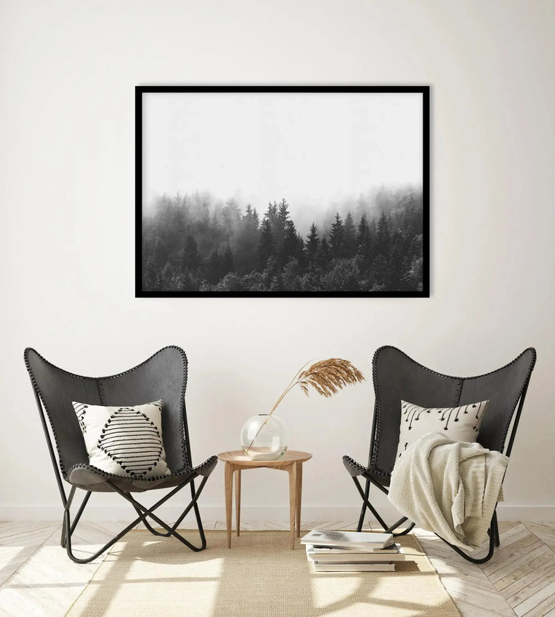 Lifestyle shot of a minimalist living room featuring a large framed art print of a misty forest in black and white. The print is displayed on a white wall above two dark grey butterfly chairs with white and black patterned cushions. A small round wooden side table with a glass vase holding dried pampas grass sits between the chairs. The room has light wooden floors and a cream rug, with natural light streaming in from the left.