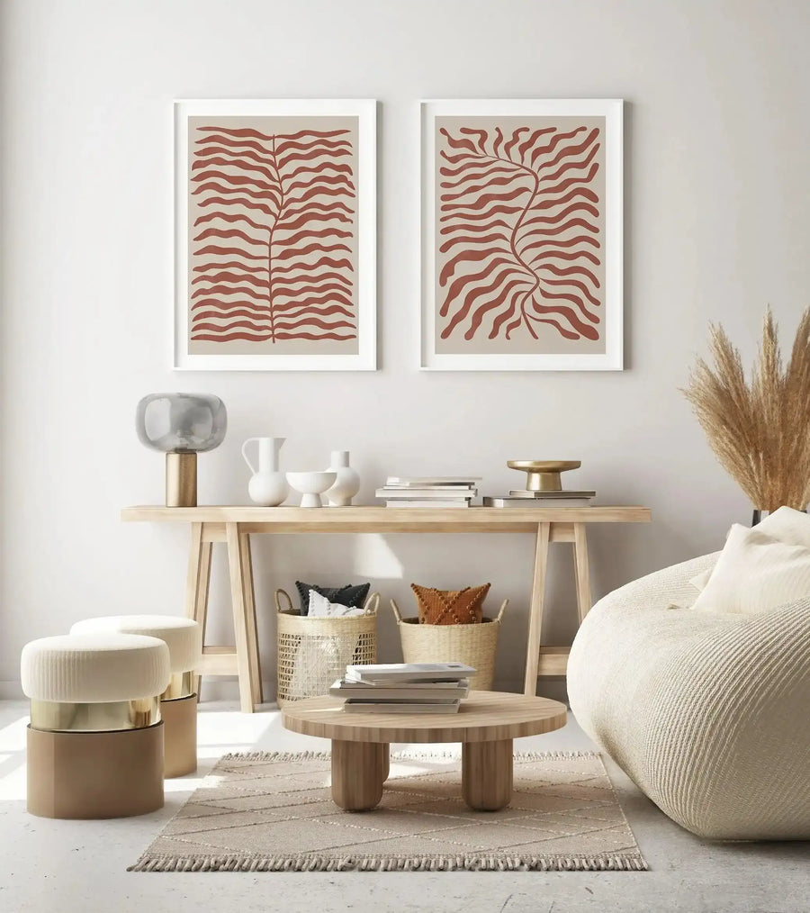 Lifestyle shot of two framed art prints displayed above a console table in a minimalist living room. The prints feature abstract botanical designs with wavy, organic leaf shapes in a reddish-brown (red earth) colour against a light beige background, with white frames and white borders. The room includes a light wood console table, a round wooden coffee table, cream-coloured ottomans, a cream textured beanbag chair, and dried pampas grass.
