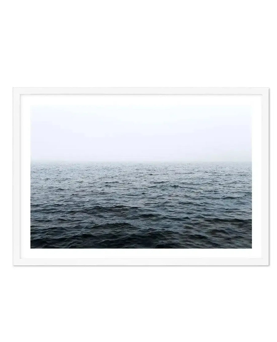 Framed photography art print of a vast, calm ocean under a misty, overcast sky, displayed in a white frame with a white border. The dark blue-grey water fills the lower two-thirds of the composition, with subtle ripples and waves, meeting a pale grey-white horizon line.