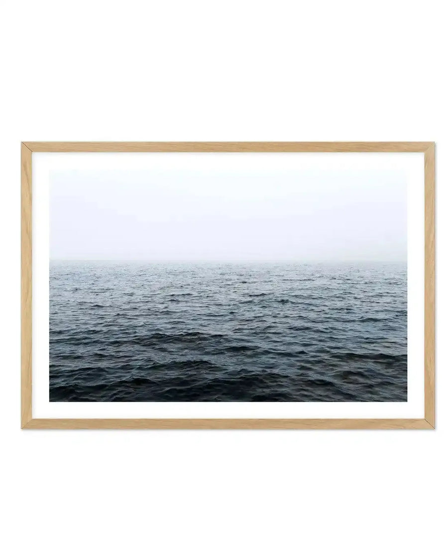 Framed photograph of a vast, calm ocean under an overcast sky, with a natural wood frame and white border. The composition features dark, deep blue-grey water with subtle ripples, meeting a hazy, light grey horizon line that blends into the pale, muted sky.