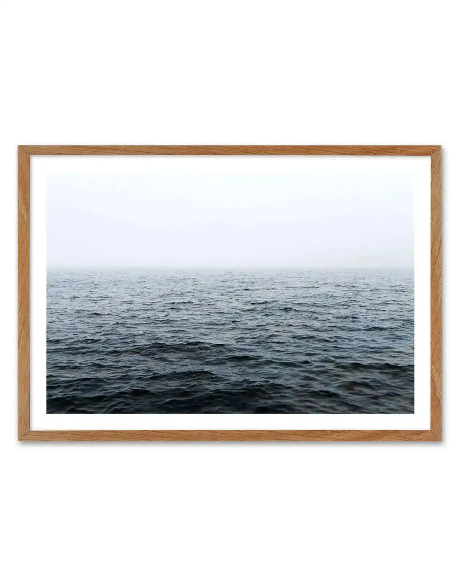 Framed photography art print of a vast, calm ocean under a misty, overcast sky, displayed in a natural oak frame with white border. The composition features dark grey-blue water with subtle ripples stretching to a hazy horizon, blending into a light grey-white sky.