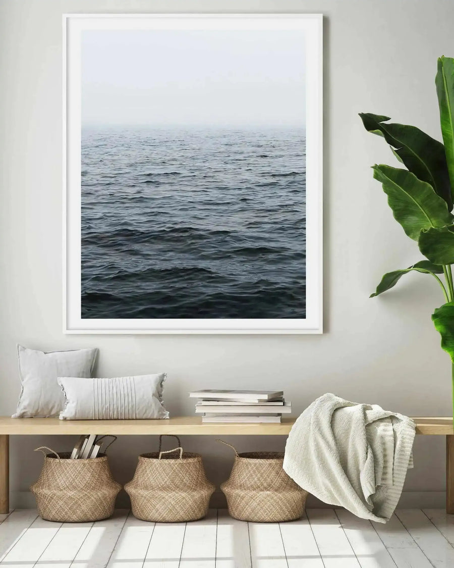 Large framed art print displayed above a wooden bench in a minimalist living room. The print features a photography of dark blue ocean waves under a misty grey sky, with a white frame and white border. The room includes two cream pillows, a stack of books, a cream throw, and three woven baskets on a white wooden floor.