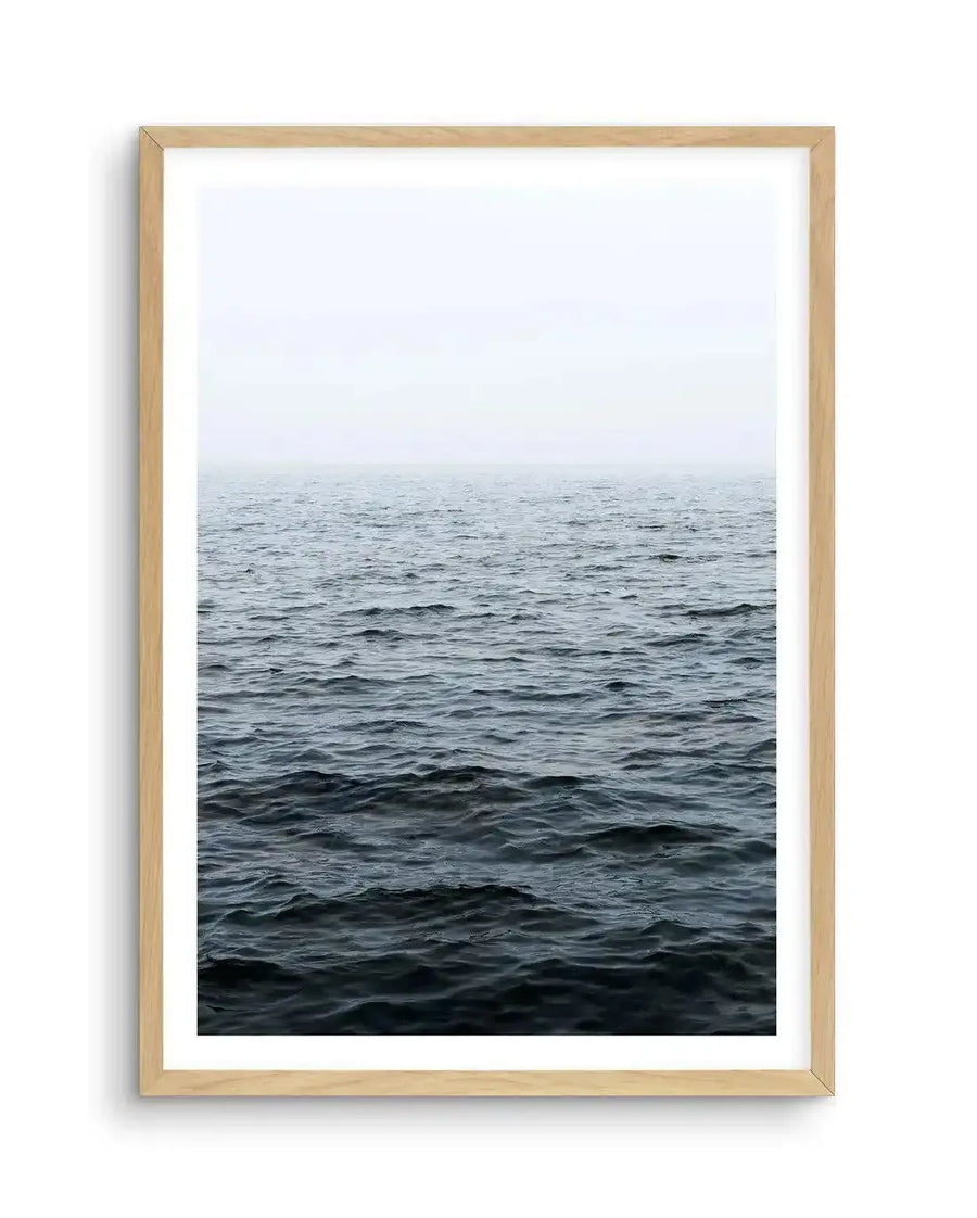 Framed photography art print of a vast ocean scene under an overcast sky, with a natural wood frame and white border. The composition features dark blue-grey water with gentle ripples stretching to a hazy horizon, meeting a pale grey-white sky.