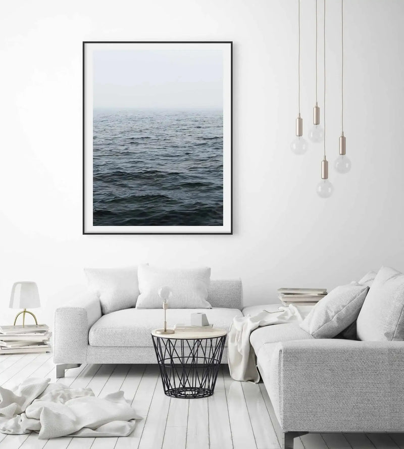 Lifestyle shot of a minimalist living room featuring a large framed art print of a moody ocean scene. The print, displayed in a black frame with a white border, depicts dark grey-blue water with subtle ripples under a hazy, light grey sky. The room includes a white sectional sofa with textured fabric, a black wire side table, a small white lamp, and several pendant lights with frosted bulbs.