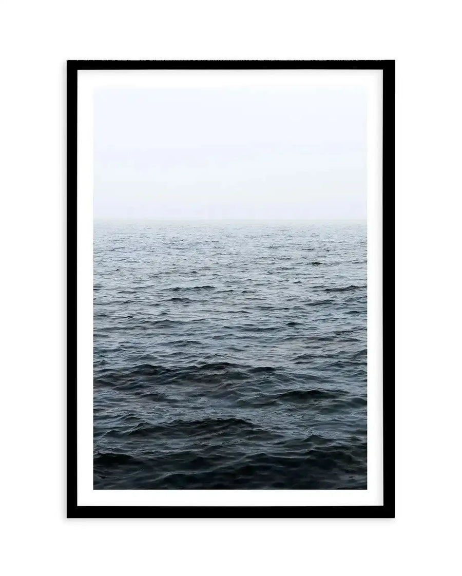 Framed photography art print featuring a minimalist seascape with a dark, choppy ocean meeting a hazy, light grey sky at the horizon, displayed in a black frame with white border.