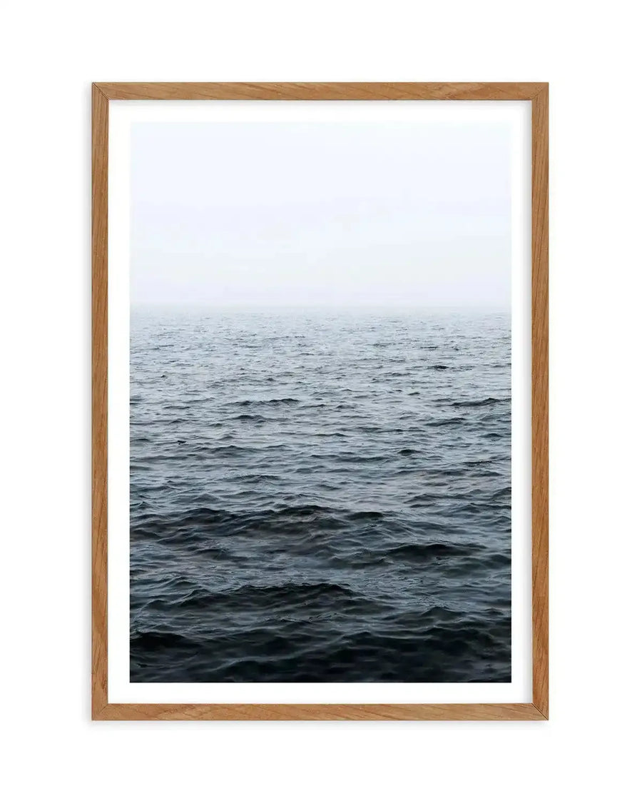 Framed photography art print of a moody ocean scene with a natural wood frame and white border. The composition features dark, choppy waves in the foreground transitioning to a lighter, misty horizon under an overcast sky.