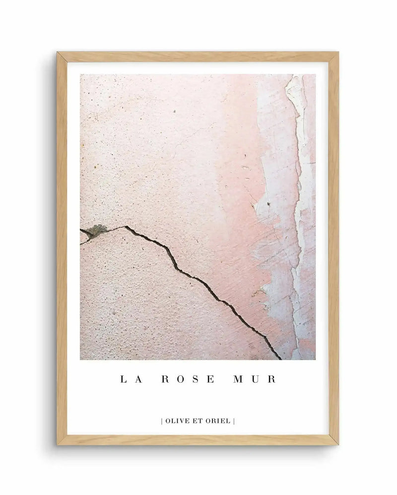 Framed art print titled "La Rose Mur" with natural wood frame and white border. The artwork features a close-up photograph of a weathered wall surface in soft dusty rose and cream tones, with peeling plaster creating an aged, textured appearance. A dark curved crack or line runs diagonally across the composition. The image has a minimalist, abstract quality capturing the subtle color gradations and worn details of the wall's surface. Text at bottom reads "OLIVE ET ORIEL" in small capitals.