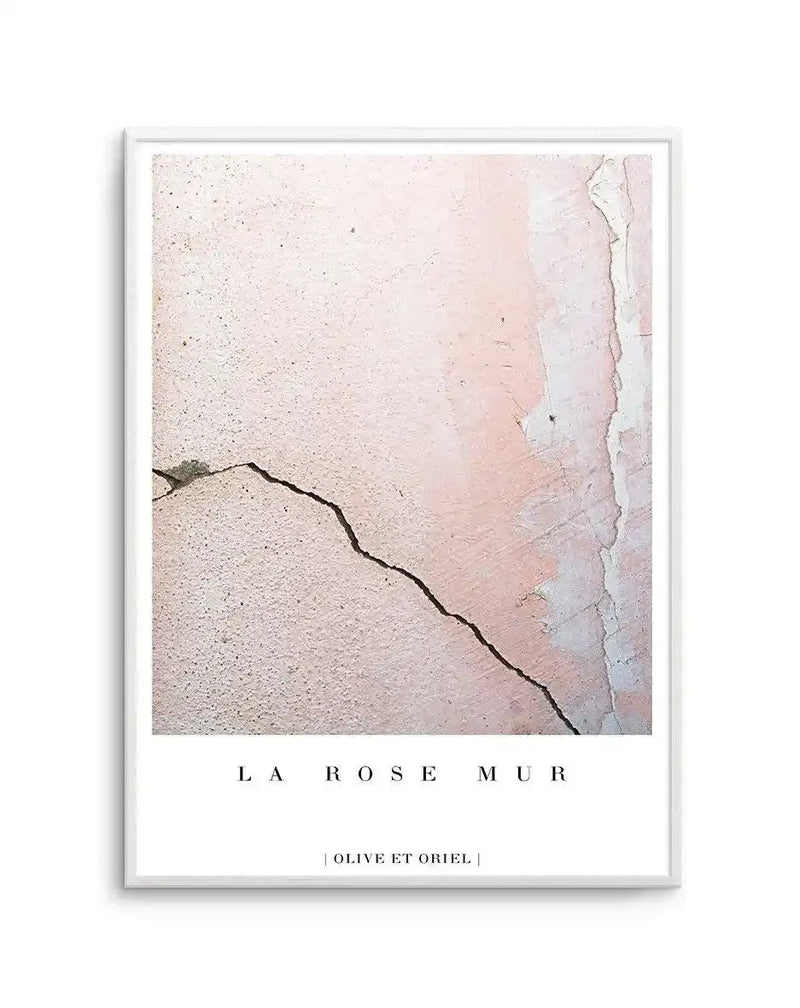 Framed art print titled "La Rose Mur" featuring a close-up photograph of a weathered pink and cream textured wall surface with a dark curved crack running diagonally across it. The wall shows peeling paint in soft dusty rose, pale pink, and off-white tones with visible wear and deterioration. A white wooden frame surrounds the image with a white border, and artist credit appears at the bottom.