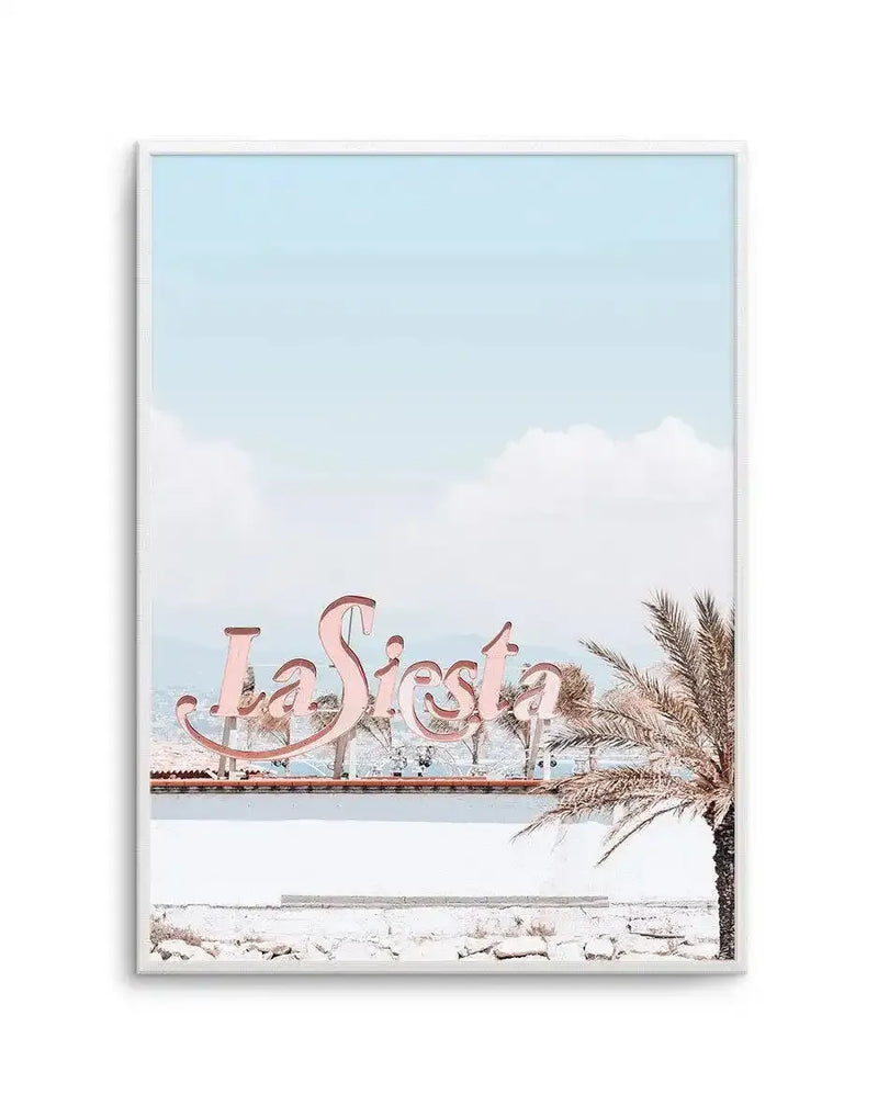 Framed photography art print featuring a retro-style pink "La Siesta" sign against a pale blue sky with fluffy white clouds, with a palm tree on the right, displayed in a white frame with white border.
