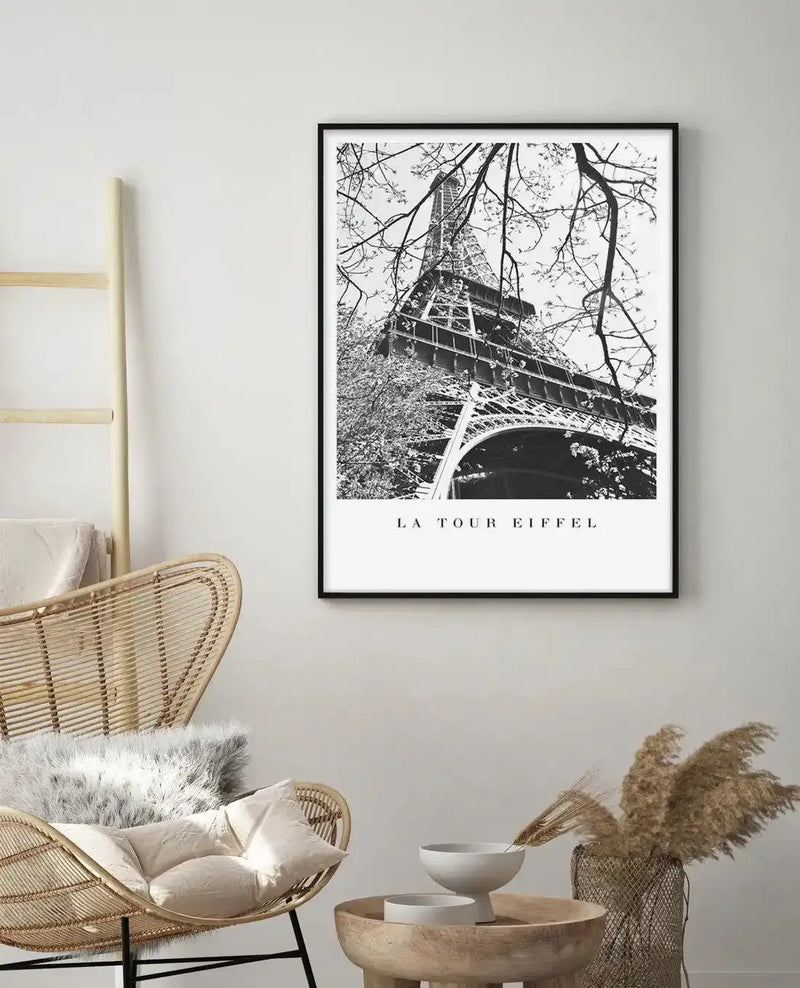 Black and white photograph of the Eiffel Tower framed by bare tree branches, printed on white background with "LA TOUR EIFFEL" text centered below the image. The print is displayed in a black frame with white border on a light gray wall above a natural wood side table and woven rattan chair with cream cushions.