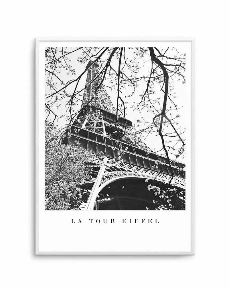 Black and white photograph of the Eiffel Tower framed by bare tree branches in the foreground, with the tower's iron lattice structure and observation decks clearly visible against a light sky. The image has high contrast and fine detail typical of vintage architectural photography. Displayed as a framed art print with the text "LA TOUR EIFFEL" centered below the photograph on a white border, mounted in a light gray frame.