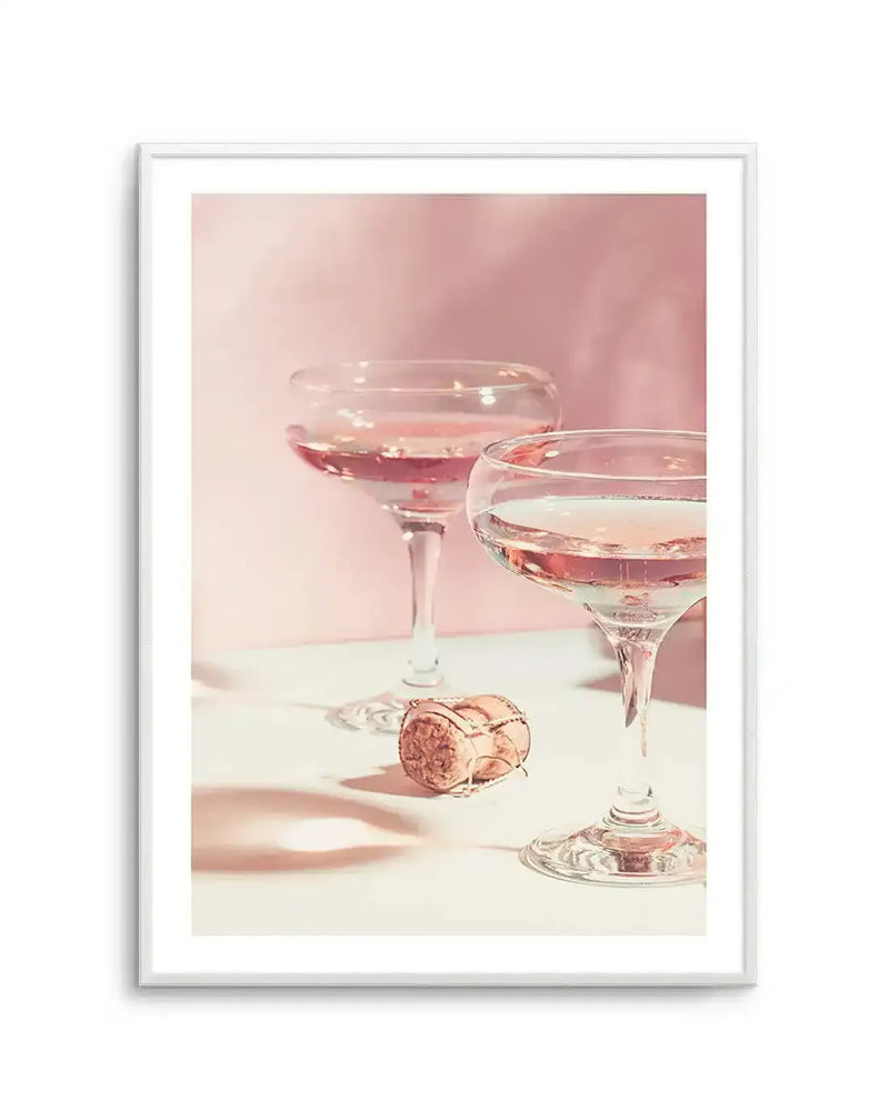 Framed photograph of two champagne coupes filled with pale pink rosé champagne, with a champagne cork lying on a white surface in the foreground. The background is a soft, diffused blush pink, creating a celebratory and elegant mood. The image is framed in a white frame with a white border.